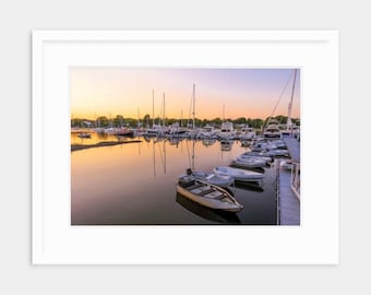 May include: A calm harbor at sunrise with a row of boats docked at a pier. The water is still and reflects the colorful sky.