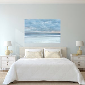 May include: A white bed with a white duvet and two white pillows with a beige pattern. Two bedside tables with lamps and a large framed photograph of a beach scene with a blue sky and white clouds above the ocean.