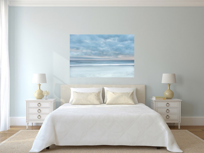 May include: A white bed with a white duvet and two white pillows with a beige pattern. Two bedside tables with lamps and a large framed photograph of a beach scene with a blue sky and white clouds above the ocean.
