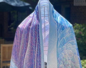 May include: A white tallit with a blue and purple abstract design. The tallit has fringes on the bottom corners and a Hebrew inscription on the left side. The tallit is displayed on a stand.