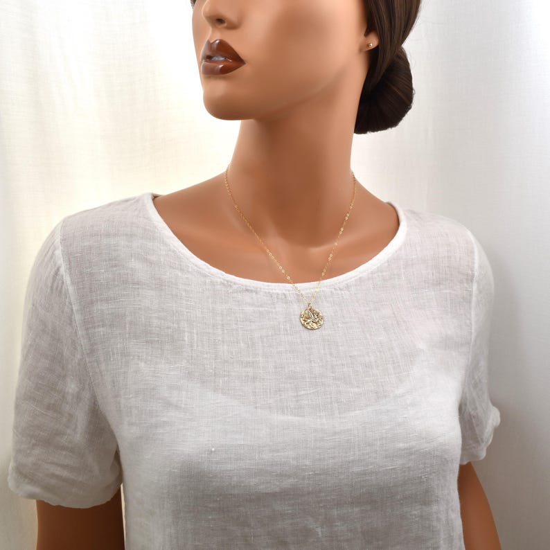 K&ouml;nnte beinhalten: Eine goldene Halskette mit einem Anh&auml;nger mit filigranem Blumendesign. Die Kette wird &uuml;ber einem wei&szlig;en Leinenoberteil getragen, was einen schlichten, aber eleganten Look erzeugt. Der Anh&auml;nger h&auml;ngt in der Mitte der Brust.