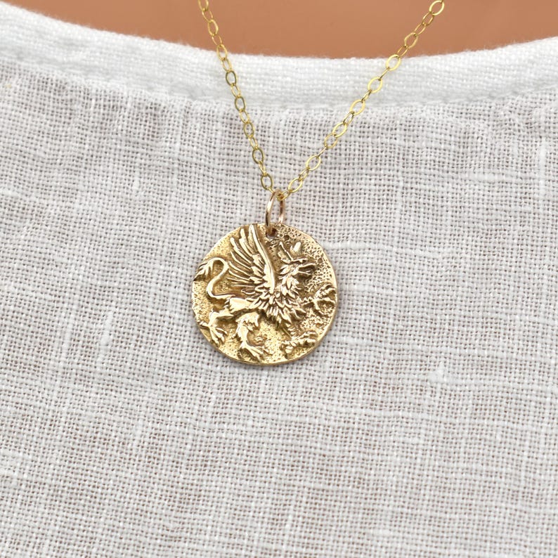K&ouml;nnte beinhalten: Goldfarbene Anh&auml;nger-Halskette mit detailliertem Greifen-Design. Der runde Anh&auml;nger h&auml;ngt an einer filigranen Kette. Der Greif, ein Fabelwesen mit Fl&uuml;geln und L&ouml;wenk&ouml;rper, symbolisiert St&auml;rke und Wachsamkeit.