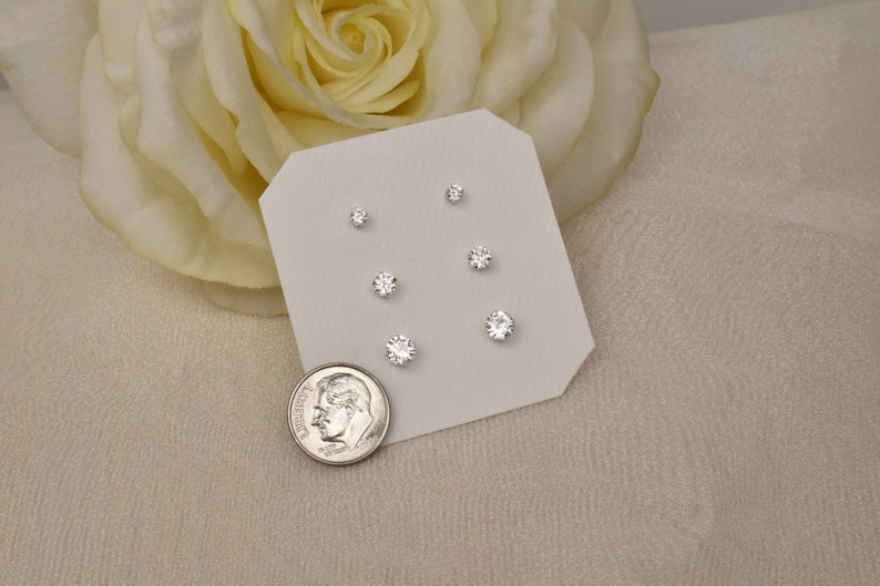 May include: A set of six silver stud earrings with clear cubic zirconia stones. The earrings are displayed on a white card with a silver dime for size comparison.
