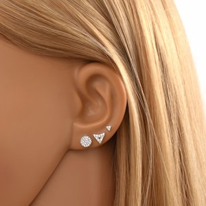 May include: Three silver stud earrings with clear cubic zirconia stones. The earrings are in the shape of a circle, a triangle, and a small dot.
