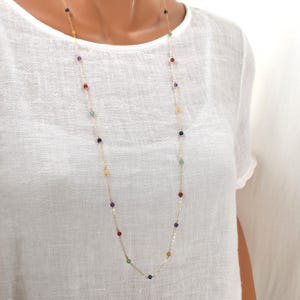 Könnte beinhalten: Eine lange goldene Halskette mit bunten Edelsteinperlen. Die Halskette hat eine zarte Kette mit gleichmäßig verteilten, kleinen, runden Perlen in verschiedenen Farben, darunter Rot, Blau, Gelb und Lila. Die Halskette wird über einem weißen Hemd getragen.