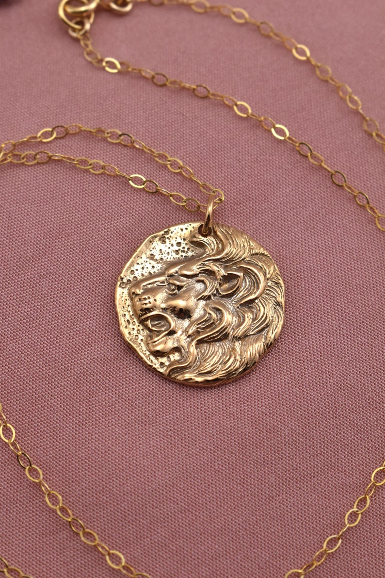 A round gold pendant with the image of a lions head is shown. The lions mouth is open as if in a roar, revealing fierce front teeth. Detailed, wavy textures depict a majestic, full mane encircling the lions head.