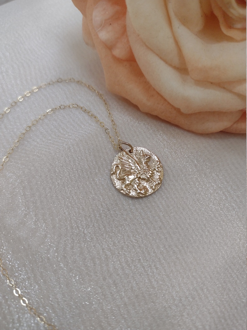 K&ouml;nnte beinhalten: Eine goldfarbene Halskette mit Anh&auml;nger, die einen strukturierten, kreisf&ouml;rmigen Anh&auml;nger mit einem detaillierten Reliefdesign aufweist. Der Anh&auml;nger h&auml;ngt an einer zarten Goldkette. Der Hintergrund umfasst einen weichen, hellfarbigen Stoff und eine pfirsichfarbene Rose.