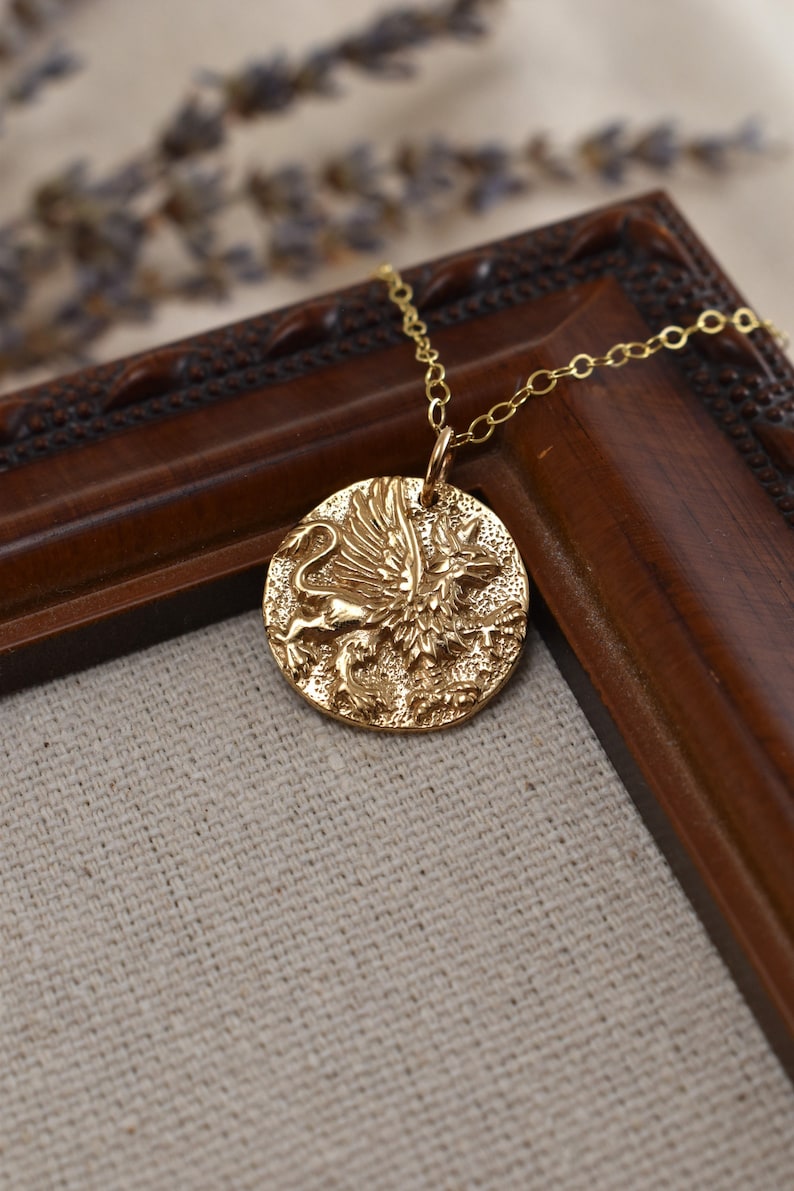 A round bronze pendant strung on a gold filled chain shows the image of a griffin, a creature from Greek mythology. It has an eagles head and talons. Feathery wings are raised proudly. It has the body of a lion, legs poised as if in mid stride.
