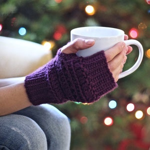May include: A person wearing purple knitted fingerless gloves is holding a white ceramic mug. The mug is filled with a warm beverage. The person is sitting in front of a Christmas tree with twinkling lights.