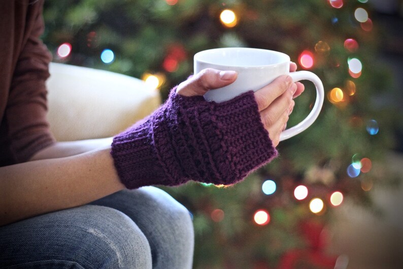 May include: A person wearing purple knitted fingerless gloves is holding a white ceramic mug. The mug is filled with a warm beverage. The person is sitting in front of a Christmas tree with twinkling lights.