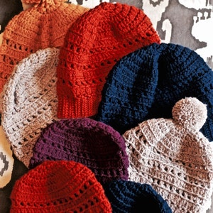 May include: A collection of eight crocheted hats in various colors, including red, orange, gray, purple, blue, and beige. The hats are arranged in a pile, with the largest hat at the bottom and the smallest hat at the top.