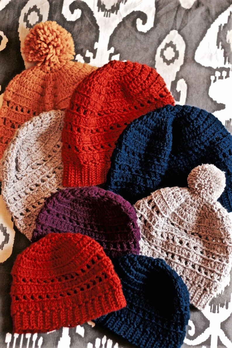 May include: A collection of eight crocheted hats in various colors, including red, orange, gray, purple, blue, and beige. The hats are arranged in a pile, with the largest hat at the bottom and the smallest hat at the top.
