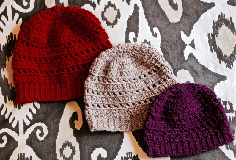 May include: Three crocheted hats in different colors: red, gray, and purple. The hats are made with a lacy stitch pattern.