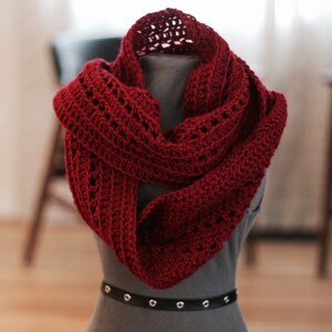 May include: A red crocheted infinity scarf with a lacy pattern. The scarf is draped over a gray mannequin.