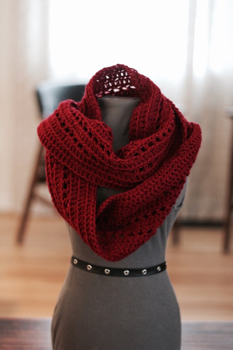 May include: A red crocheted infinity scarf with a lacy pattern. The scarf is draped over a gray mannequin.