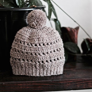 May include: A crocheted beanie hat in a light brown color with a fluffy pom pom on top. The hat has a textured stitch pattern and is sitting on a dark wooden surface.
