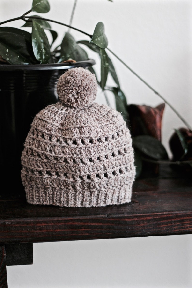 May include: A crocheted beanie hat in a light brown color with a fluffy pom pom on top. The hat has a textured stitch pattern and is sitting on a dark wooden surface.