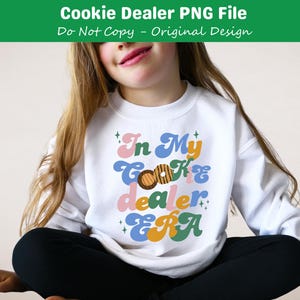 May include: A white sweatshirt with the text "In My Cookie Dealer Era" in colorful retro-style lettering. The text is surrounded by small stars and a cookie graphic.