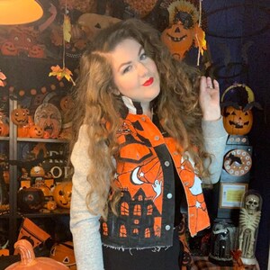 Puede incluir: Una chaqueta vaquera negra recortada con un diseño pintado a mano de una casa encantada, un fantasma y una luna creciente. La chaqueta tiene detalles en naranja y negro y mangas grises.