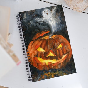 Puede incluir: Una libreta de espiral negra y naranja con una pintura de una calabaza de Halloween con un fantasma flotando sobre ella.