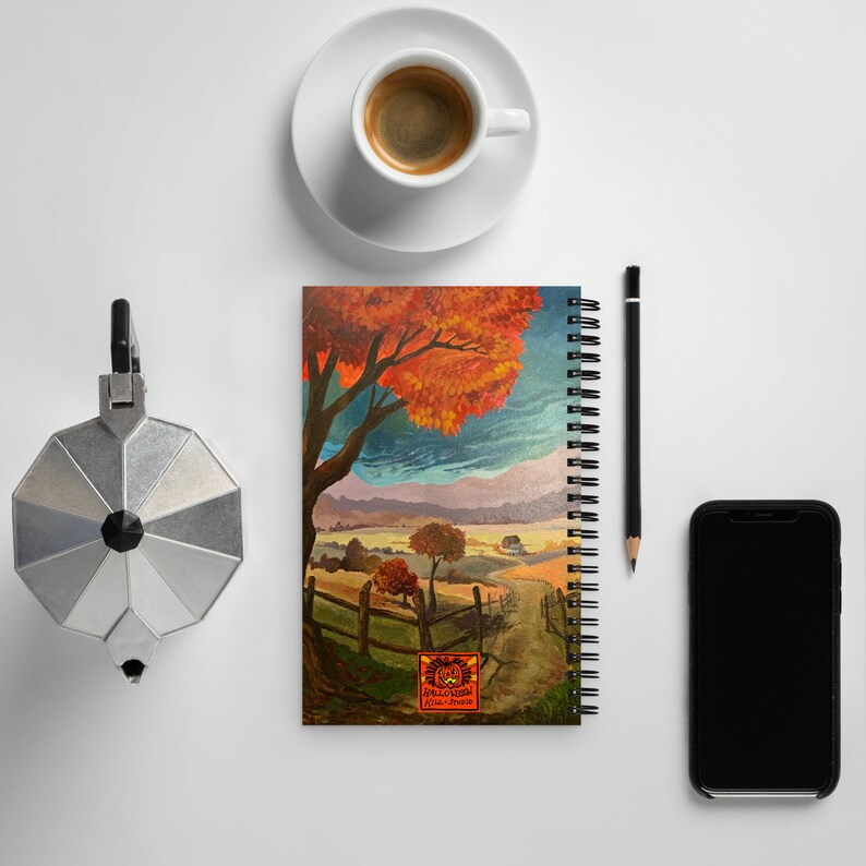 May include: A spiral bound notebook with a colorful autumn landscape illustration. The cover features a large tree with orange and red leaves, a winding path, and a small wooden fence. The text "Halloween Hill Studio" is printed on the bottom of the cover.