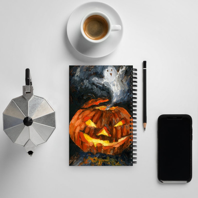 Puede incluir: Un cuaderno de espiral con una imagen pintada de una calabaza de Halloween con un fantasma que emerge de la parte superior. El fondo es azul oscuro con pinceladas negras y blancas.