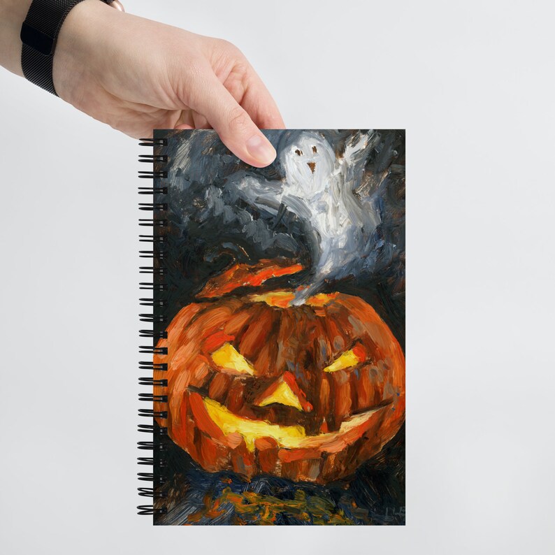 Puede incluir: Un cuaderno de espiral con una pintura de una calabaza de Halloween con un fantasma que se eleva desde la parte superior. La calabaza es naranja y tiene una cara tallada con una amplia sonrisa y ojos brillantes. El fantasma es blanco y tiene una leve sonrisa.