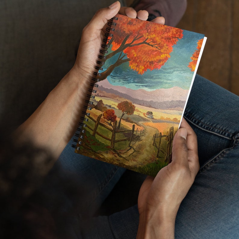 May include: A spiral bound sketchbook with a colorful painting of a fall landscape. The painting features a large orange tree, a winding path, and a distant mountain range.