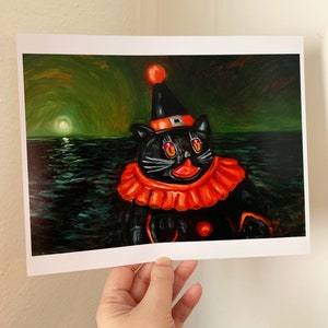 Op de afbeelding: Een zwarte kat met een rode en oranje narrenmuts en kostuum, met een rode neus en grote ogen, drijft in een donkergroene zee met een heldere maan op de achtergrond.