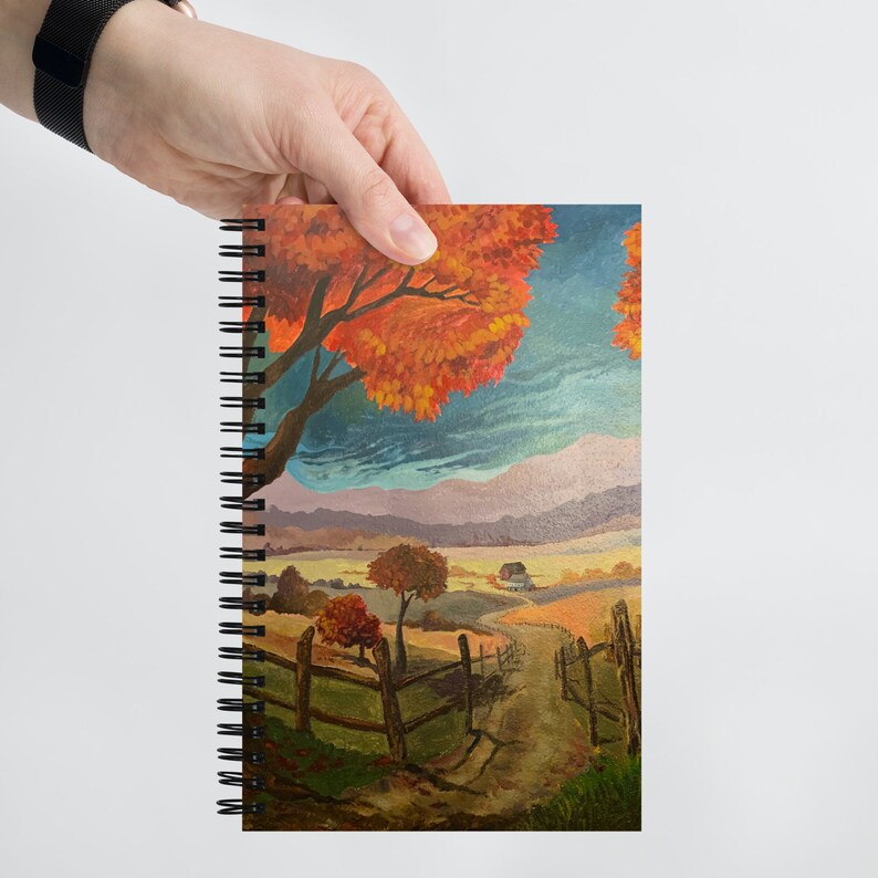 May include: A spiral bound notebook with a colorful painting of a rural landscape. The painting features a winding path leading to a small house in the distance, with trees in the foreground and a blue sky with clouds above.