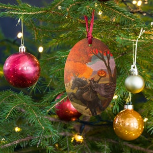 Puede incluir: Adorno navideño ovalado de madera con una escena del Jinete sin Cabeza con una cabeza de calabaza sobre un caballo negro en un paisaje otoñal. El adorno cuelga de una cinta roja en un árbol de Navidad con adornos rojos y dorados.