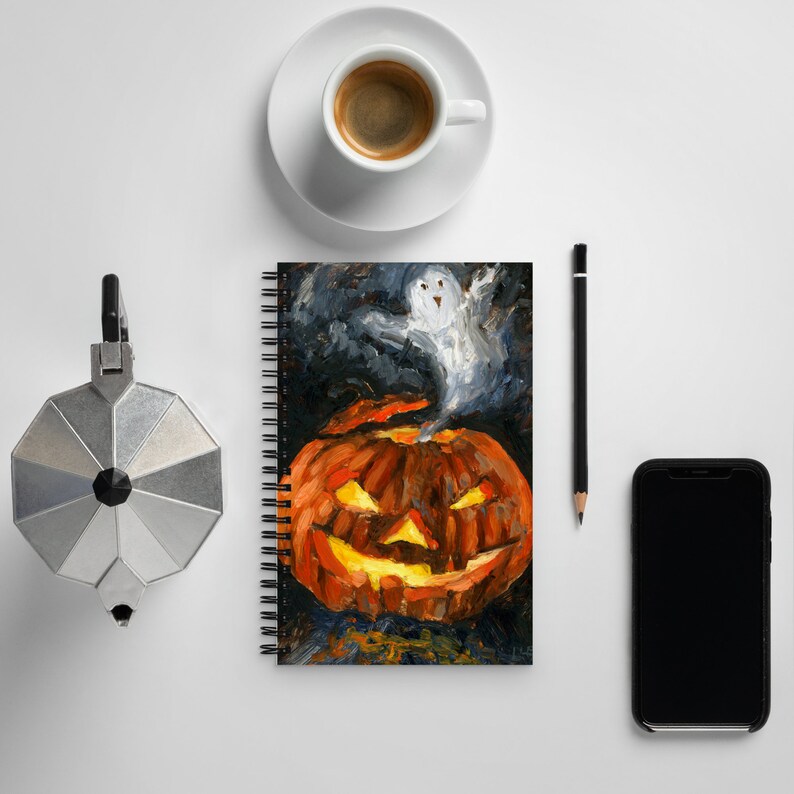 Puede incluir: Una libreta de espiral con fondo negro y una pintura de una calabaza de Halloween con un fantasma volando desde la parte superior. La calabaza es naranja y tiene una cara de miedo.