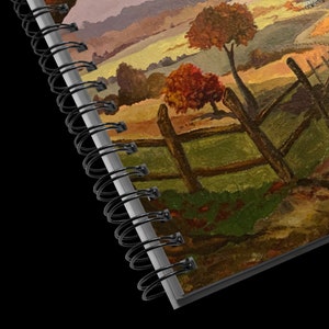 May include: A close-up of a spiral bound notebook with a painted image of a rural landscape. The image features a wooden fence, trees with fall foliage, and a distant farmhouse.