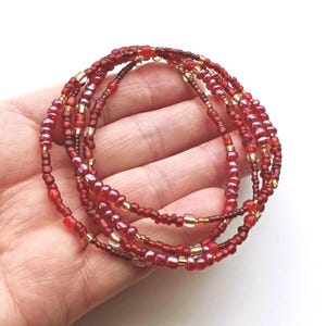 May include: A multi-strand beaded bracelet featuring a collection of small, round glass beads in various shades of deep red, crimson, and burgundy, interspersed with occasional clear and metallic gold-toned accent beads strung on a flexible cord.