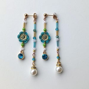 Puede incluir: Un par de pendientes colgantes con cuentas azules y verdes, un dije de flor azul y una perla en la parte inferior.
