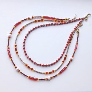 Puede incluir: Tres hileras de collares de cuentas con cuentas rojas y doradas y detalles de perlas blancas.