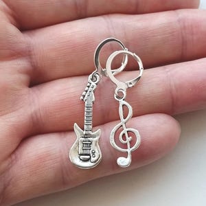 Op de afbeelding: Een paar zilverkleurige oorbellen met muzikale bedels. De ene oorbel is een gedetailleerde hanger in de vorm van een elektrische gitaar en de andere is een vioolsleutel. Beide bedels hangen aan kleine, ronde, scharnierende metalen ringen.