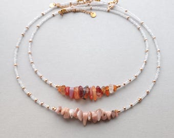 May include: Two layered necklaces featuring small clear glass beads interspersed with tiny gold-toned metal beads. The top necklace displays a cluster of polished, irregular-shaped orange and red stone chips, while the bottom necklace features a cluster of pale pink stone chips.