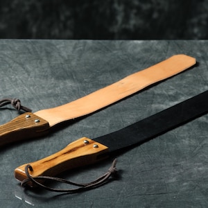 May include: Two leather paddles with wooden handles and leather straps. One paddle is brown leather and the other is black leather.