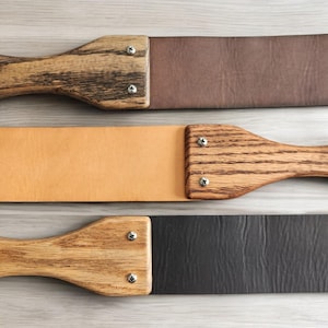 May include: Three leather strop tools with wooden handles. The strops are brown, tan, and black. The handles are made of wood with a grain pattern.