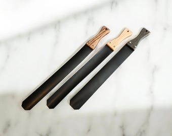 以下が含まれることがあります： 木製のハンドルが付いた3本の長く平らな黒いスパチュラ。各ハンドルには明るい色から暗い色まで独特の木目模様があり、2つの小さな金属製リベットで黒いブレードに固定されています。各ハンドルには小さな穴が開いています。