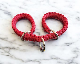 May include: A bright red paracord accessory featuring two circular loops connected by a central braided section. Three polished silver-toned metal rings are attached to the structure, with one ring positioned between the two loops and two rings at the base of each loop.