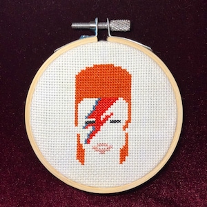 May include: A cross-stitch embroidery of David Bowie's face with his signature lightning bolt makeup. The embroidery is framed in a wooden hoop.