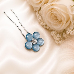 Può includere: Una forcina color argento con un ornamento a forma di fiore a cinque petali. I petali sono realizzati in materiale azzurro semitraslucido con un leggero luccichio e un cristallo trasparente sfaccettato è incastonato al centro del fiore.