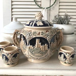 Puede incluir: Un juego de jarras de cerveza de cerámica vintage con una jarra grande con tapa y pequeñas tazas a juego. La jarra presenta una escena de castillo, racimos de uvas y texto en alemán. El juego es beige, azul y marrón.