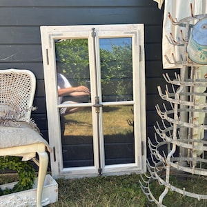 Könnte beinhalten: Ein rechteckiger Fensterrahmen mit abgenutzter weißer Oberfläche und zwei Glasscheiben. Der Rahmen verfügt über einen zentralen Metallverschluss und Metallscharniere an den Seiten. Das Glas spiegelt die Umgebung und das Grün wider.