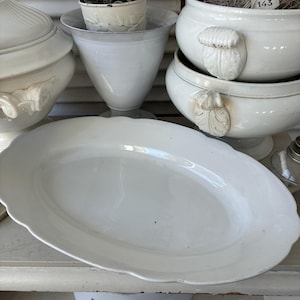 May include: A collection of white ceramic serving dishes and decorative pots. The centerpiece is a large, oval platter with a scalloped edge. Other items include lidded tureens and a pedestal vase, all in a vintage style.