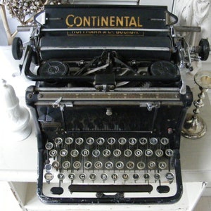 May include: A black vintage Continental typewriter with a gold inscription. The typewriter has a standard QWERTY keyboard and a carriage return lever.