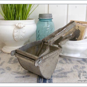 May include: An antique, metal potato ricer with a perforated basket and hinged handles. The ricer is displayed with a white ceramic planter, a blue glass jar, and vintage photographs, evoking a vintage aesthetic.