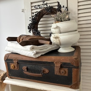 Op de afbeelding: Een zwarte en bruine vintage koffer met metalen sluitingen, met daarop een stapel witte stof, een houten klos met touw, een krans van donkere bessen en twee gestapelde witte keramische kommen met decoratieve handvatten en bloemen.